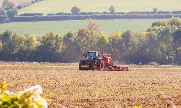 Green tractor pulling machinery across a field, with fields and hedgerows in the background.